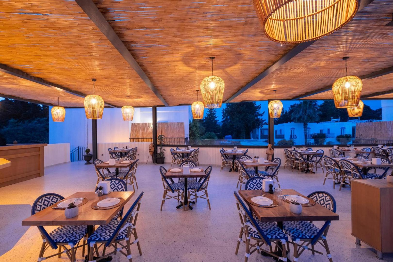 Paloma Marina Suites restorāna terase ar stilīgiem pītiem gaismekļiem, koka galdiem un zili baltiem krēsliem vakara stundā