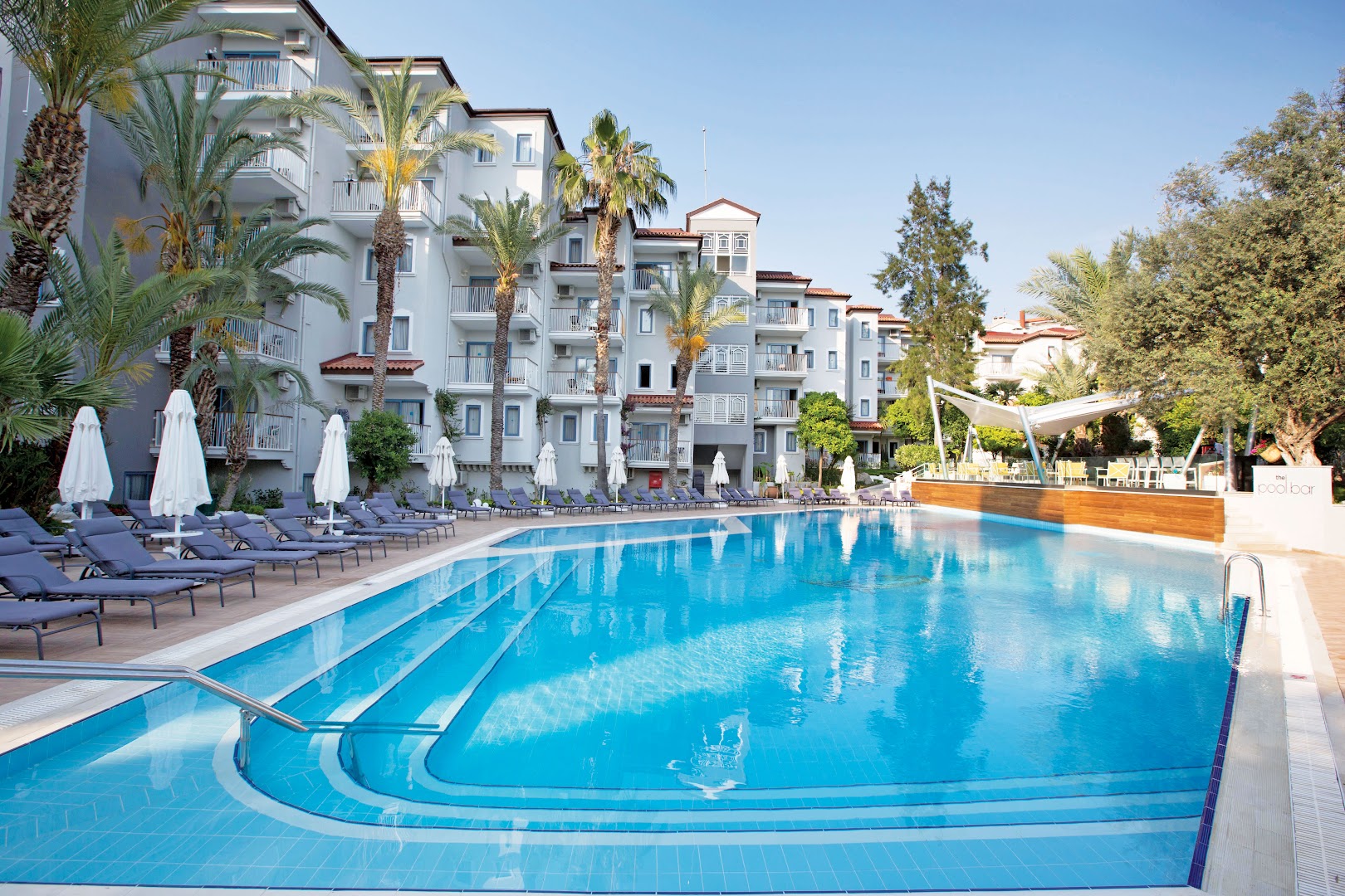 Paloma Marina Suites āra baseins ar ziliem flīžu pakāpieniem un sauļošanās krēsliem Kušadasi kūrortā Turcijā, ko ieskauj palmas un viesnīcas korpusi.