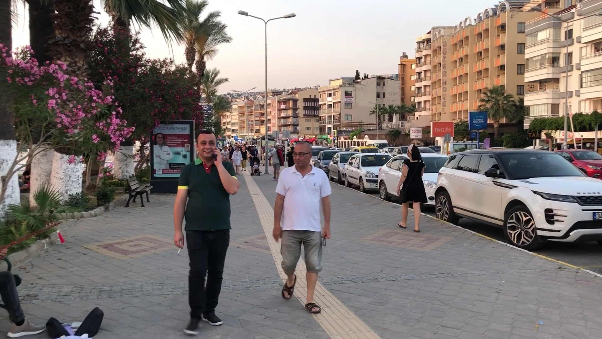 Kušadasi pilsētas promenāde pie Paloma Marina Suites viesnīcas — gājēju iela ar palmām, veikaliem un automašīnām piekrastes zonā