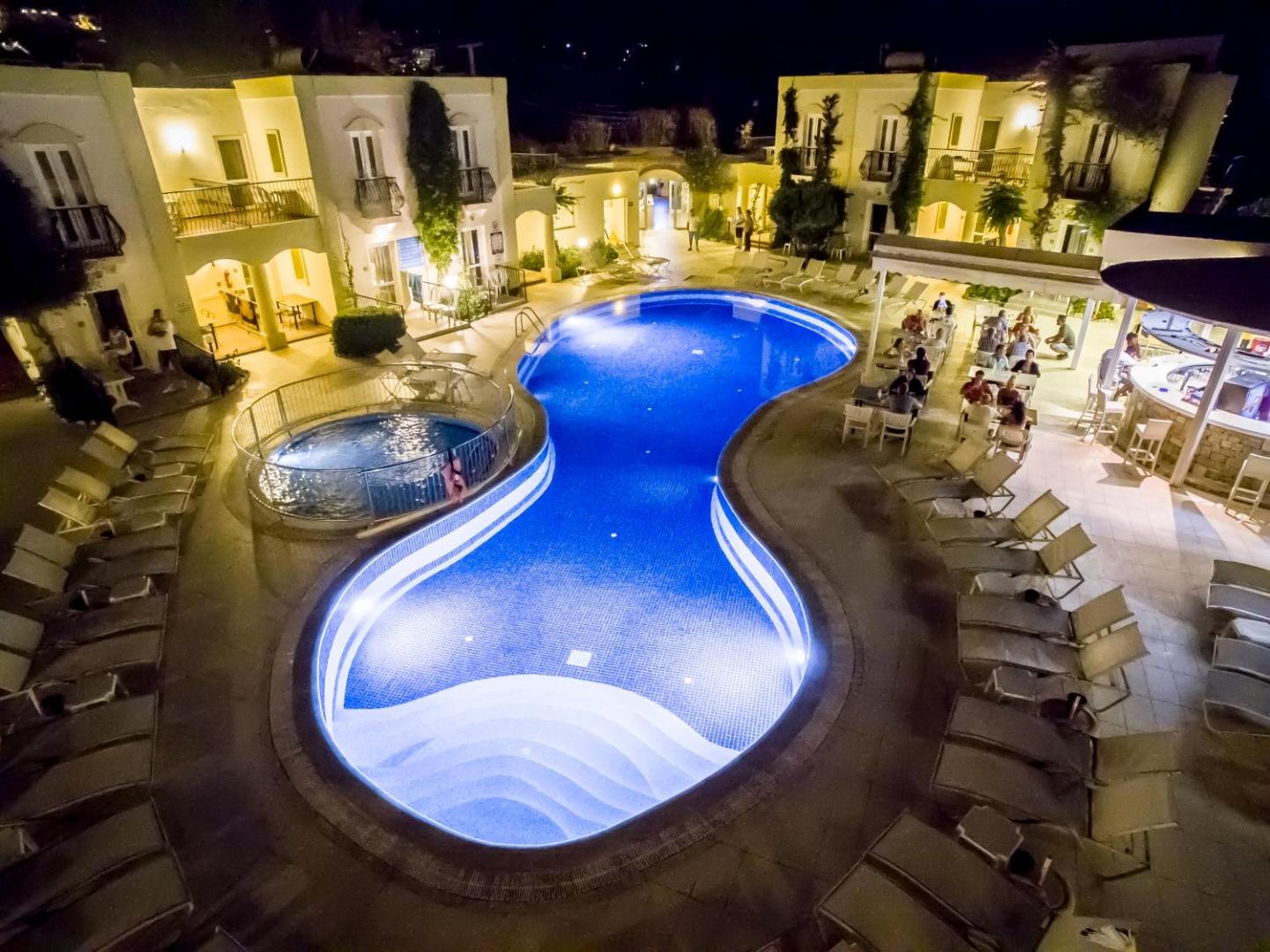 Paloma Marina Suites izgaismots baseins nakts laikā ar mirdzošām gaismām un viesiem, kuri bauda vakariņas āra terasē pie bāra.
