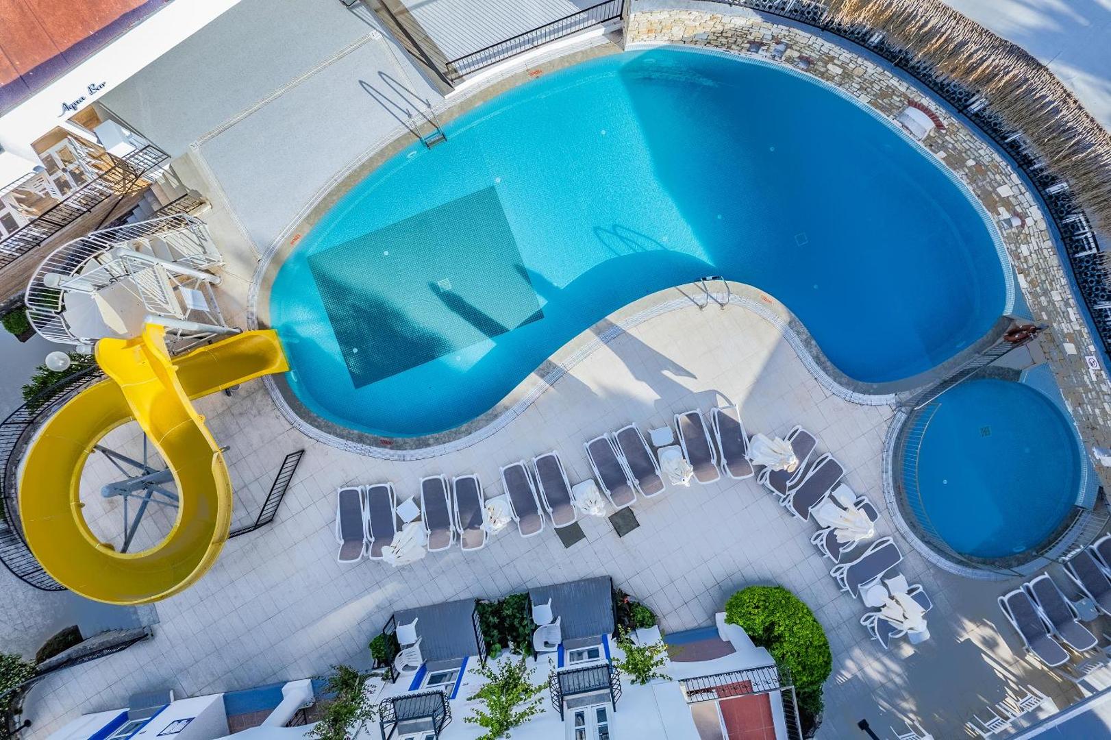 Skats no augšas uz Paloma Marina Suites baseinu ar dzeltenu ūdens slidkalniņu, bērnu baseinu un sauļošanās krēsliem pie Aqua Bar.