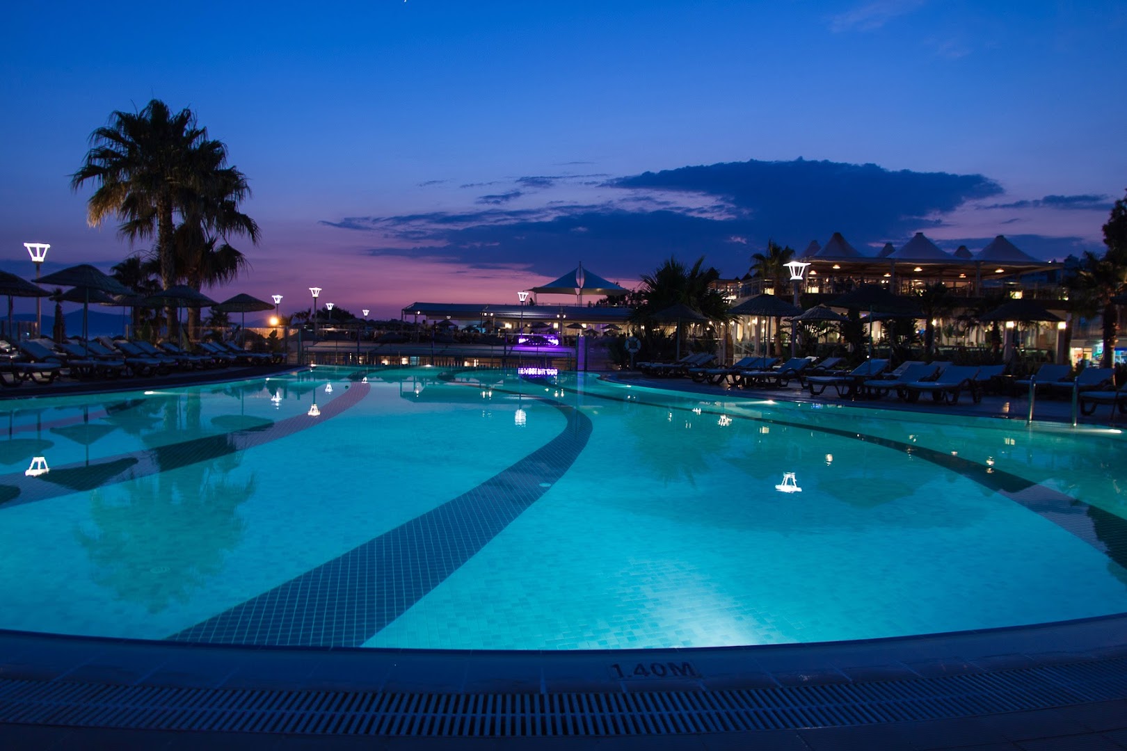 Armonia Holiday Village & Spa izgaismots baseins vakarā ar palmām un saulrieta debesīm fonā Bodrumas reģionā.