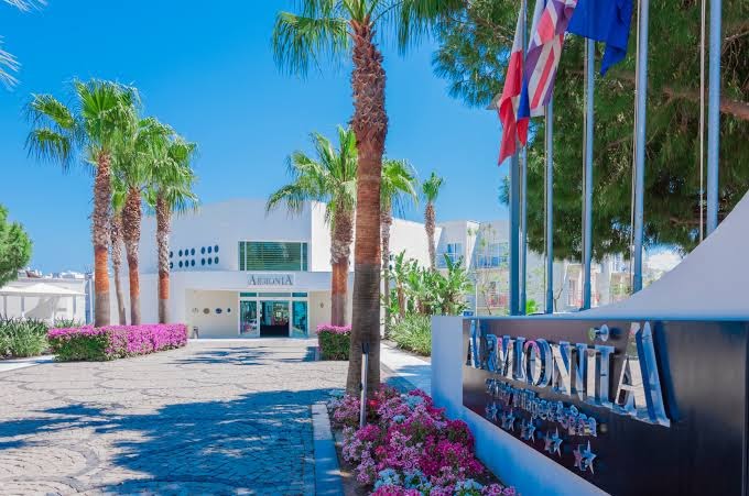 Armonia Holiday Village & Spa galvenā ieeja ar viesnīcas nosaukumu, palmām un karogiem pie ieejas mezgla