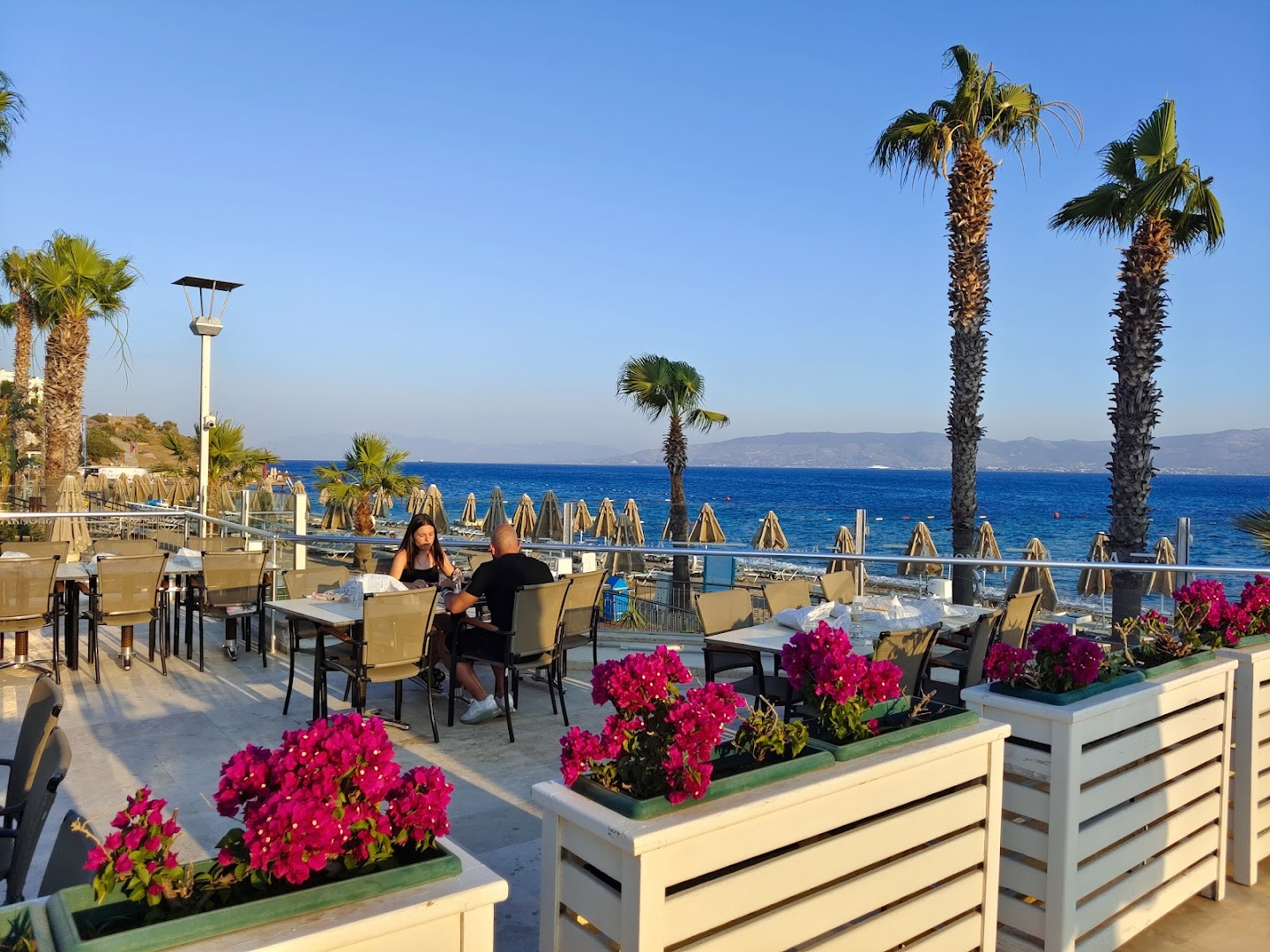Armonia Holiday Village & Spa āra terase ar galdiņiem, palmām un ziedošām bugenvillijām, skats uz jūru saulrietā