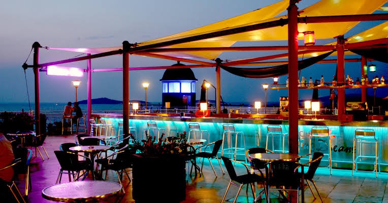 Vakara atmosfēra Blue Dreams Resort & Spa āra bārā ar neona apgaismojumu, galdiņiem un skatu uz jūru saulrietā