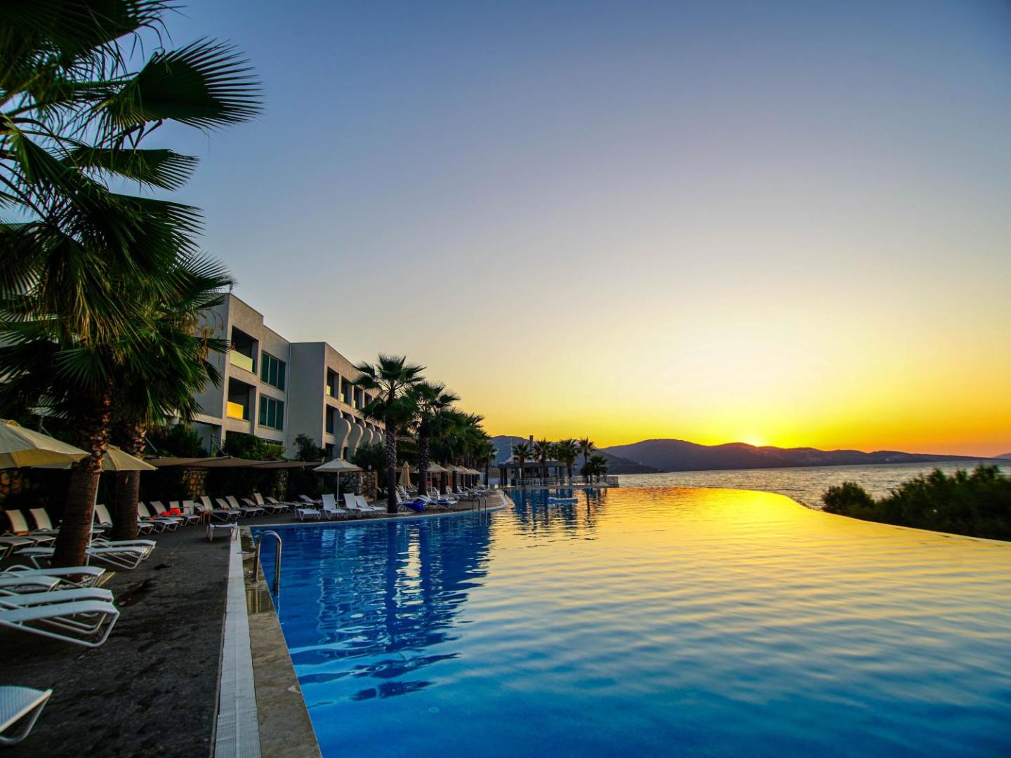 Saulriets pie Blue Dreams Resort & Spa bezmalas baseina — debesis un ūdens virsma krāsojas zeltainos toņos pie viesnīcas korpusa.