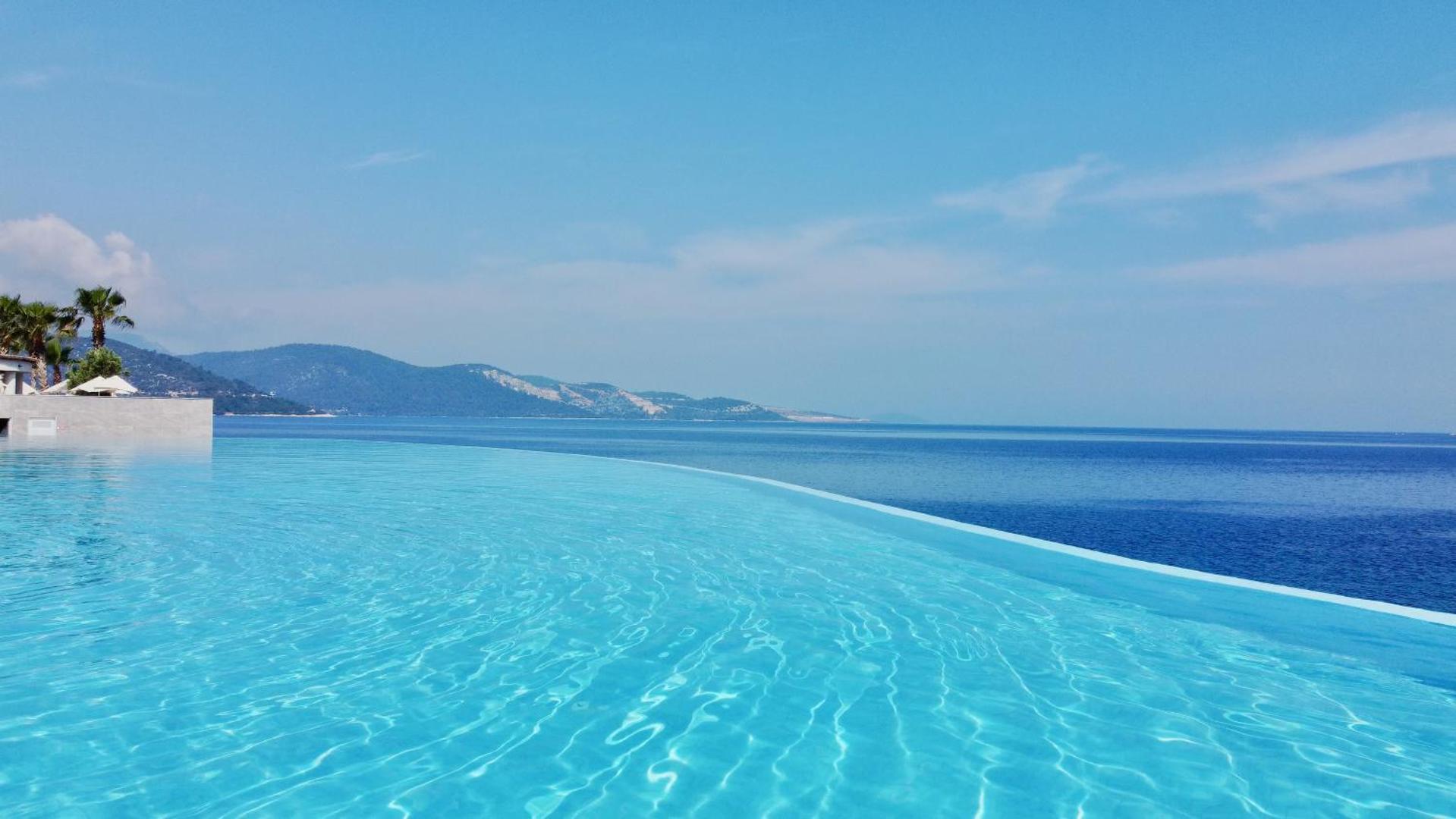 Tuvplāns Blue Dreams Resort & Spa bezmalas baseina ūdenim, kas saplūst ar zilo Egejas jūru un kalnu ainavu tālumā.