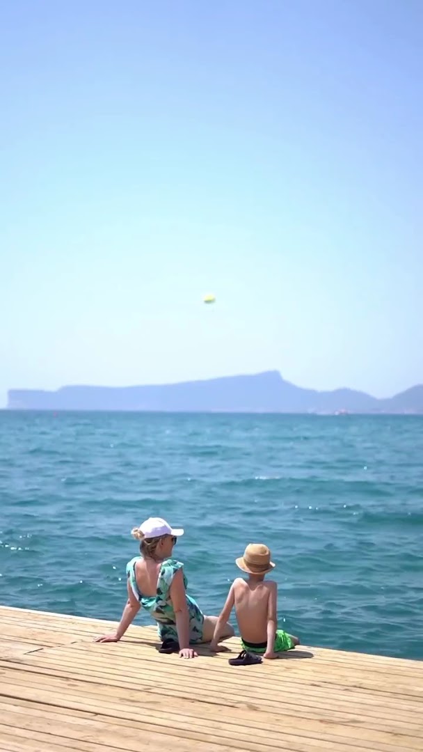 Māte ar bērnu sēž uz Ulusoy Kemer Holiday Club koka piestātnes un vēro Vidusjūras panorāmu Kemeras piekrastē