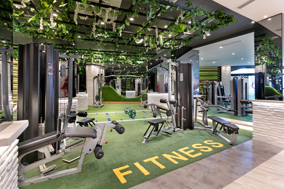 Limak Arcadia Sport Resort moderns fitness centrs ar trenažieriem un mākslīgā zālāja segumu, dekorēts ar zaļumiem pie griestiem