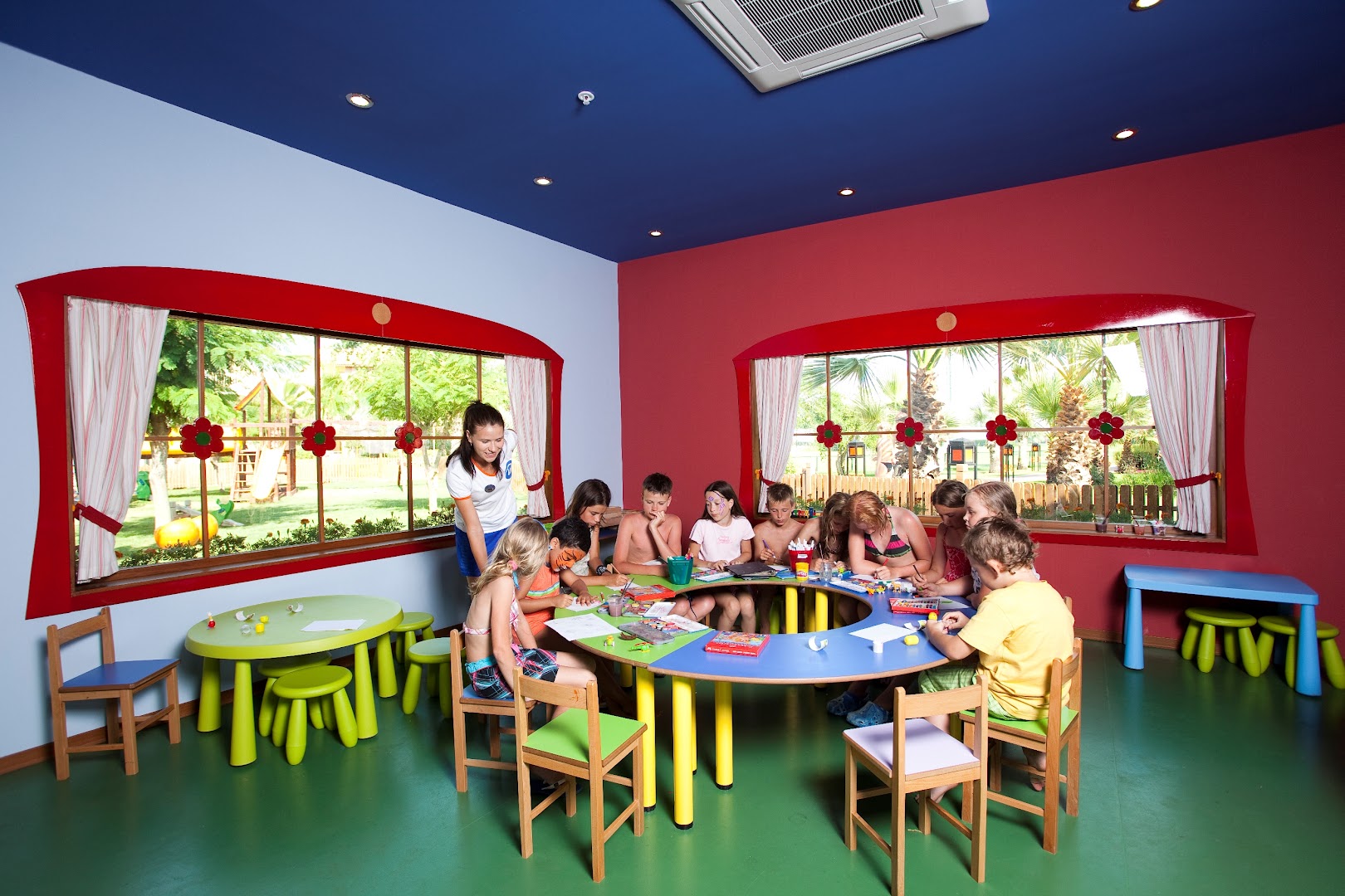 Limak Arcadia Sport Resort bērnu klubs ar krāsainām mēbelēm un bērniem, kuri zīmē pie apaļa galda pedagoga uzraudzībā