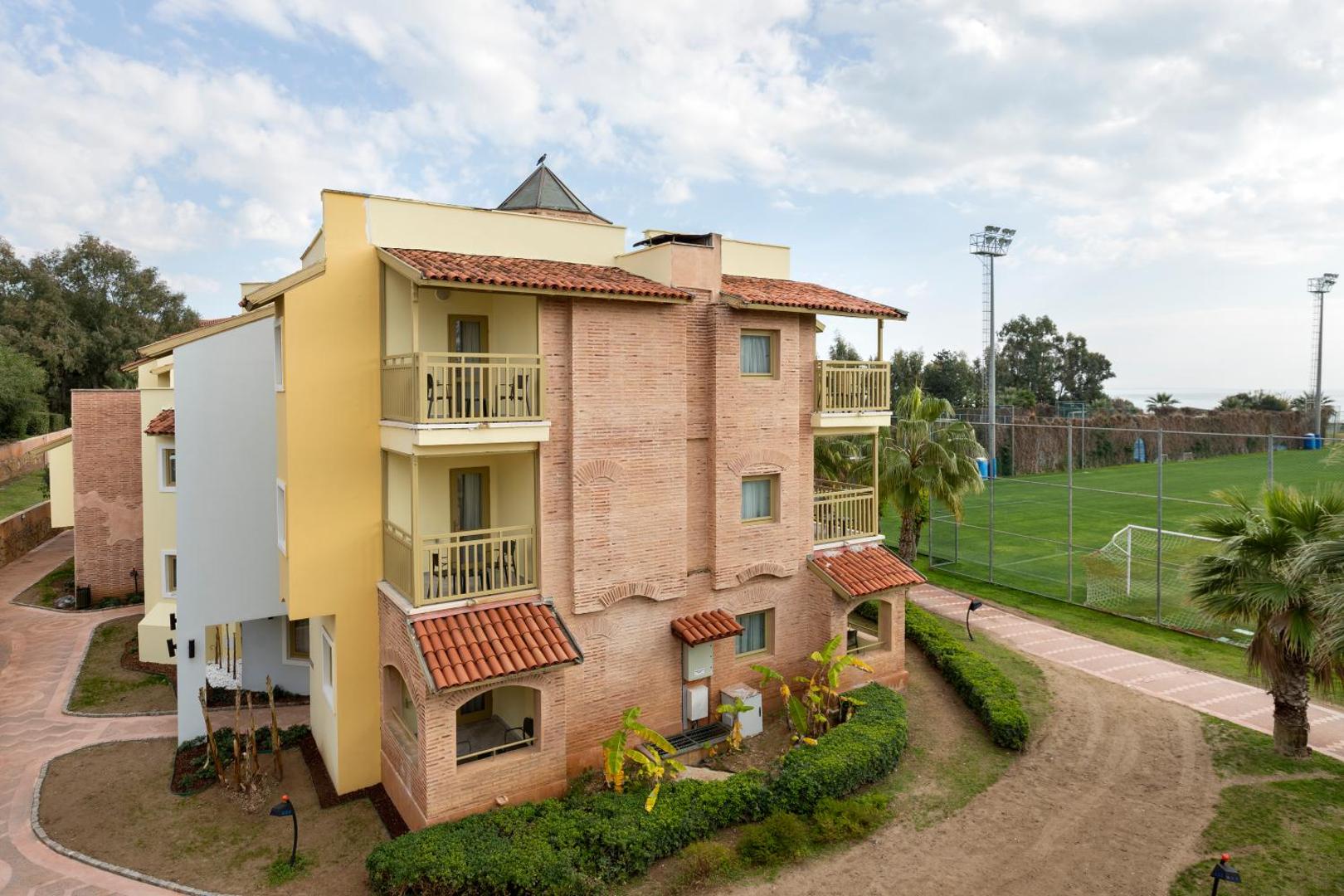 Limak Arcadia Sport Resort dzīvojamais korpuss ar balkoniem blakus futbola laukumam — ķieģeļu un dzeltenas krāsas fasāde Belekas kūrortā