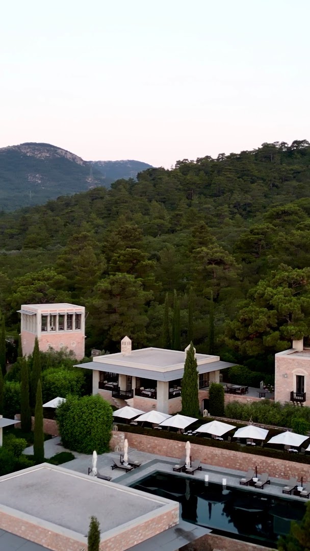 Skats no augšas uz Amanruya Resort teritoriju priežu meža ieskāvumā — baseina zona ar saulessargiem un kalnu panorāmu fonā