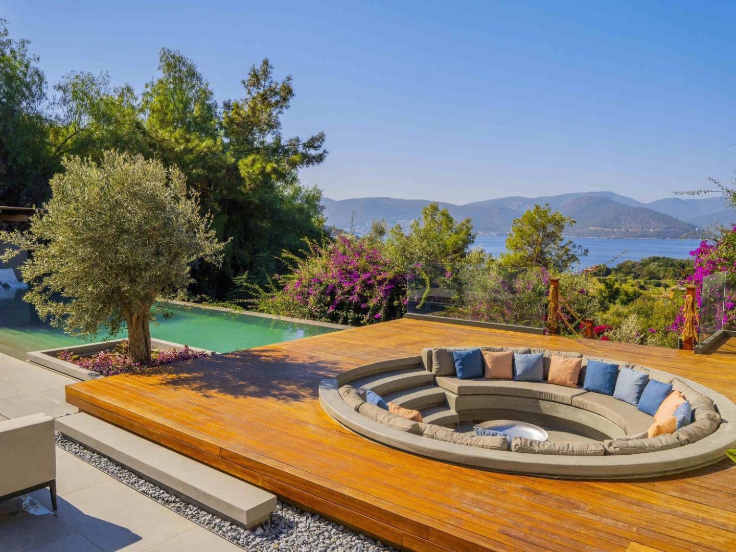 Rixos Premium Bodrum terase ar iebūvētu atpūtas zonu, spilveniem, olīvkokiem un skatu uz zilo jūras līci un kalniem