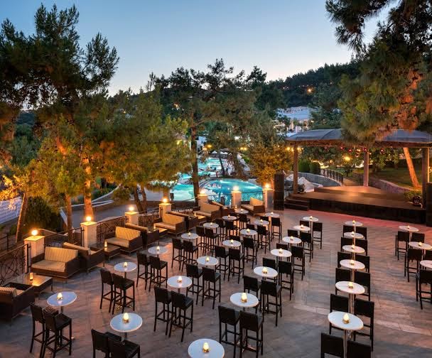 Rixos Premium Bodrum āra terase ar bāra galdiņiem, skatuvi un apgaismotu baseinu priežu ieskautā teritorijā vakarā