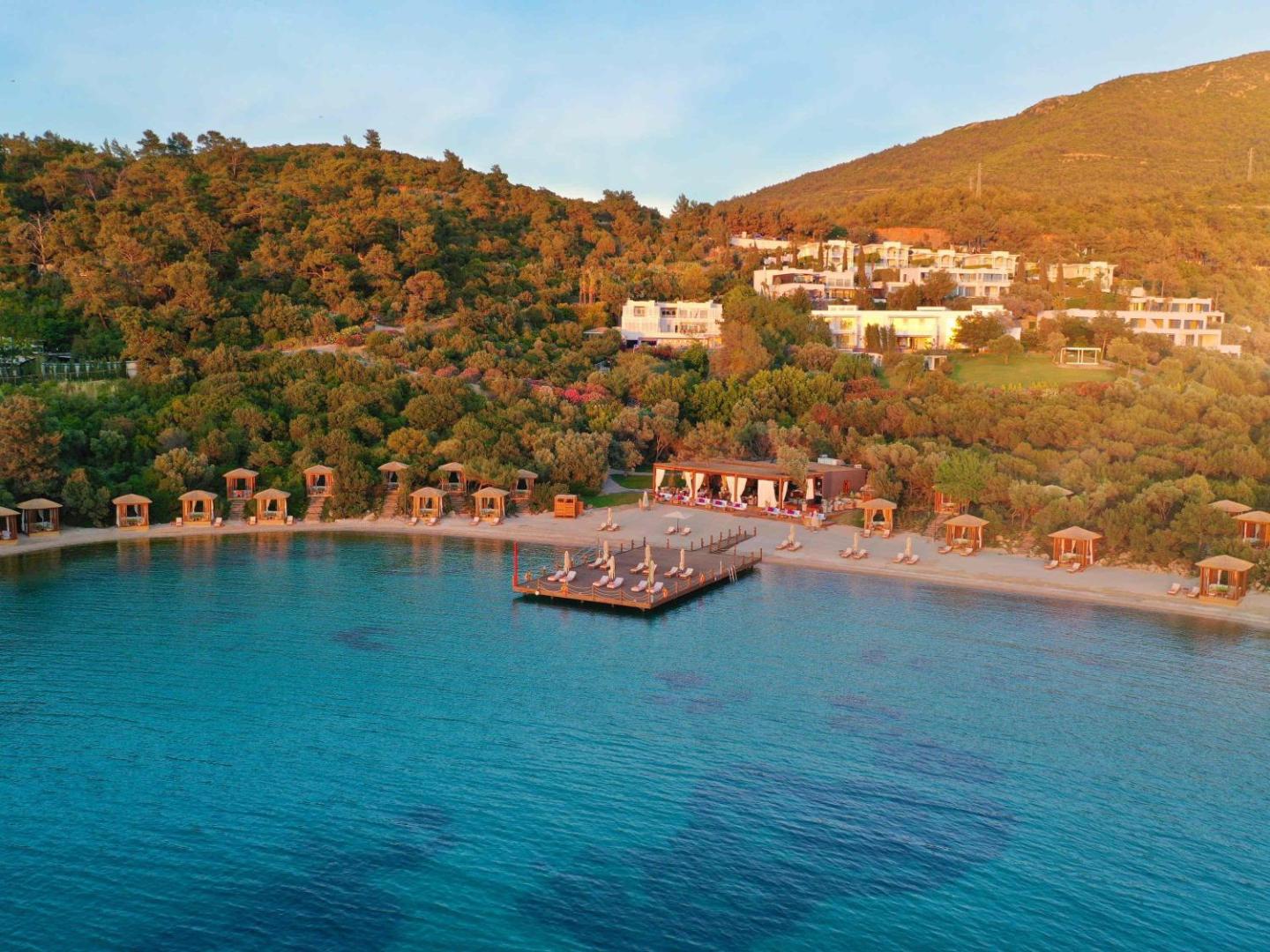 Rixos Premium Bodrum bungalo un pludmales zona saulrieta gaismā — kalnu ieskauta viesnīcas teritorija pie līča