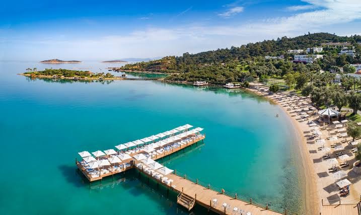Rixos Premium Bodrum privātā pludmale ar koka laipu, sauļošanās krēsliem un saulessargiem dzidrā jūras līcī Bodrumā