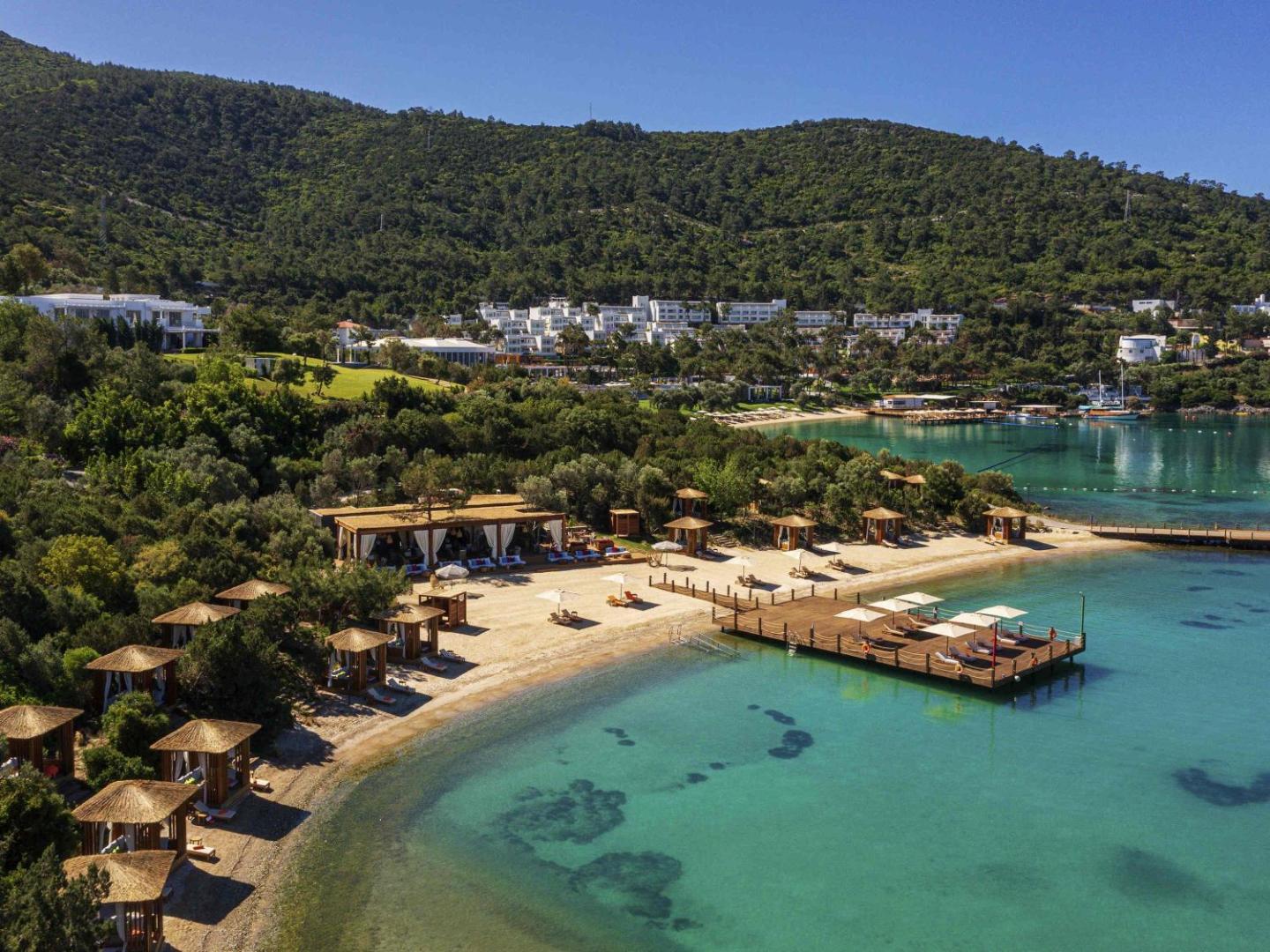 Rixos Premium Bodrum panorāmas skats uz pludmali, koka platformu un viesnīcas ēkām kalna nogāzē zaļumu ieskāvumā