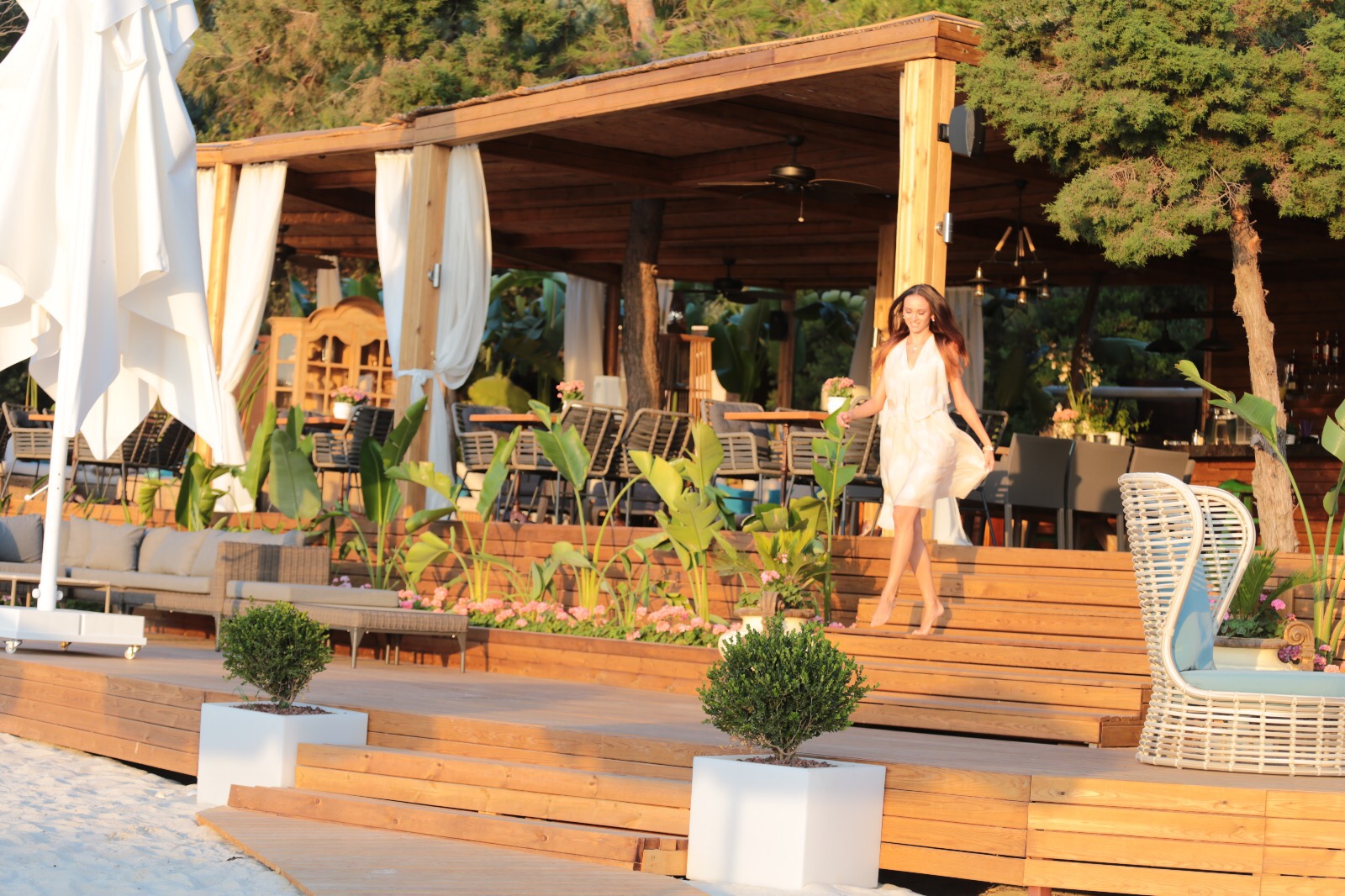Rixos Premium Bodrum pludmales bārs un atpūtas zona ar koka konstrukcijām, dīvāniem un sievieti baltā kleitā