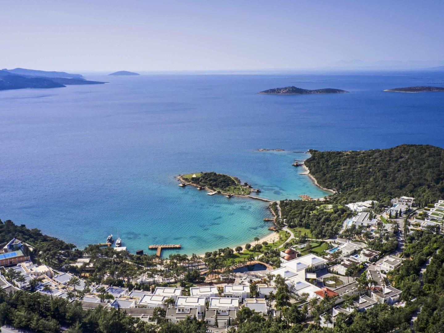 Rixos Premium Bodrum viesnīcas panorāmas skats no putna lidojuma uz Egejas jūras līci, balto villu kompleksu un apkārtējiem zaļajiem pakalniem