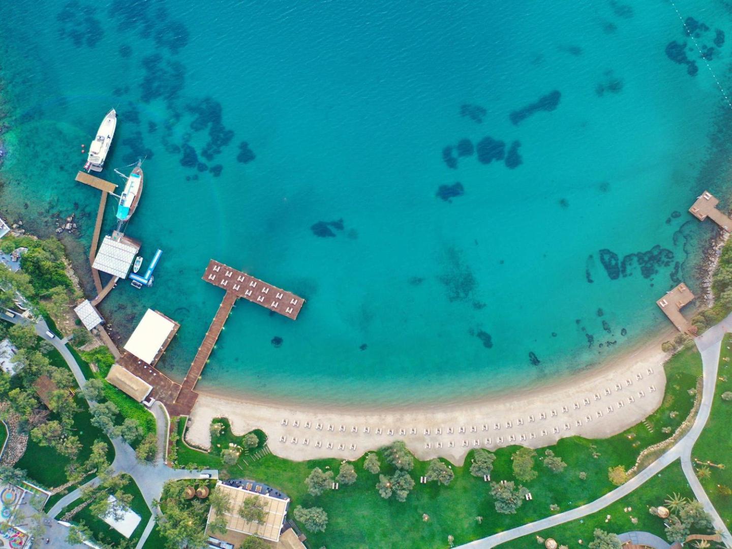 Rixos Premium Bodrum privātā pludmale no putna lidojuma — tirkīza krāsas ūdens, smilšu josla ar saulessargiem un koka laipas