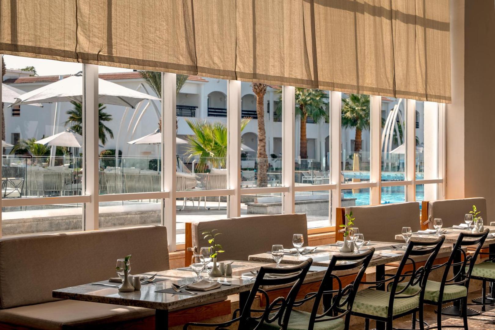 Jaz Sharm Dreams Resort restorāna iekštelpas ar klātiem galdiem pie lieliem logiem, no kuriem paveras skats uz kūrorta teritoriju Šarm eš Šeihā.