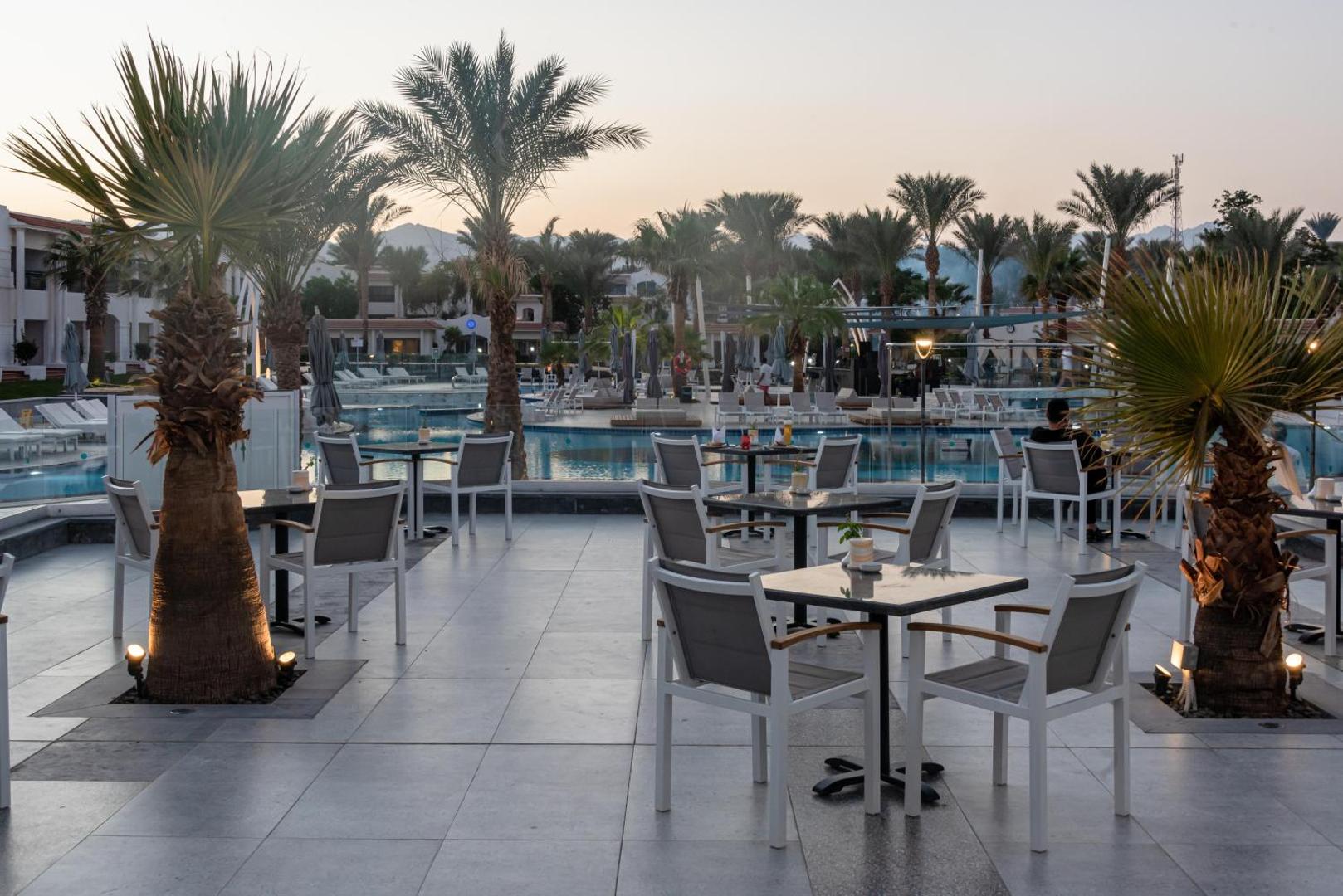 Jaz Sharm Dreams Resort āra terase ar galdiņiem un krēsliem pie baseina saulrieta laikā Šarm eš Šeihā, apkārtnē palmas un kalni.