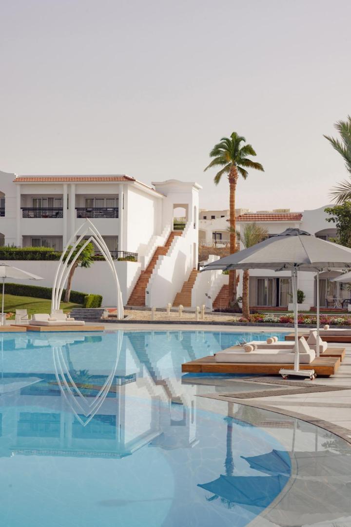 Jaz Sharm Dreams Resort viesnīcas korpuss pie baseina ar baltām kāpnēm, sauļošanās krēsliem un palmām saulainā dienā Ēģiptē