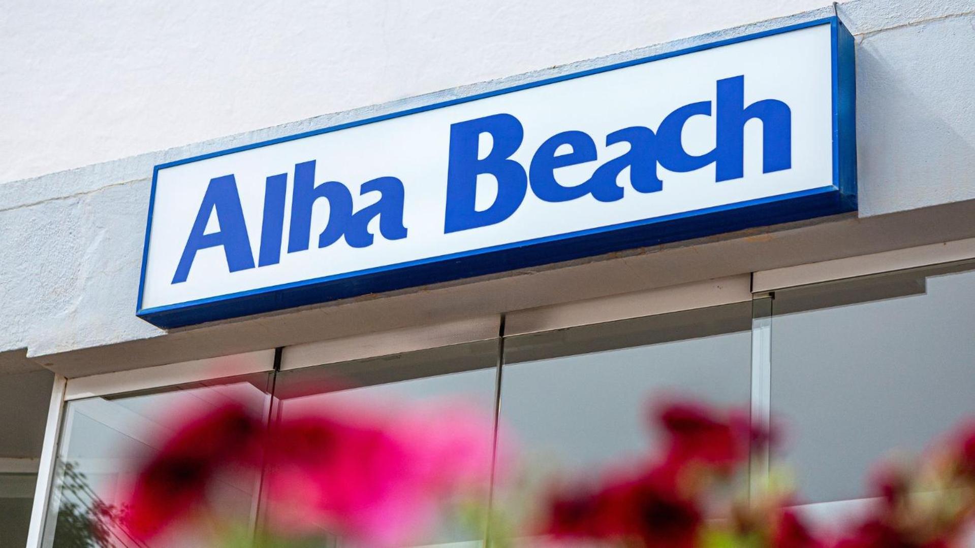 MedPlaya Hotel Alba Beach viesnīcas nosaukuma izkārtne zilā krāsā uz balta fona virs stikla durvīm ar ziediem priekšplānā
