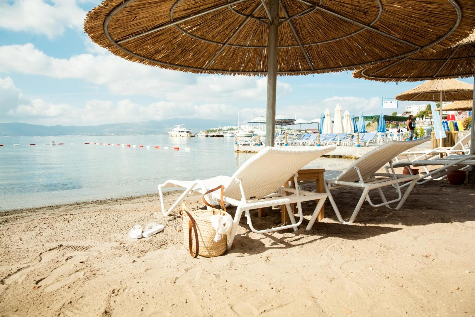Sentido Bellazure privātā pludmale Bodrumā ar baltiem sauļošanās krēsliem zem salmu saulessargiem smilšainā krastā pie dzidra jūras ūdens