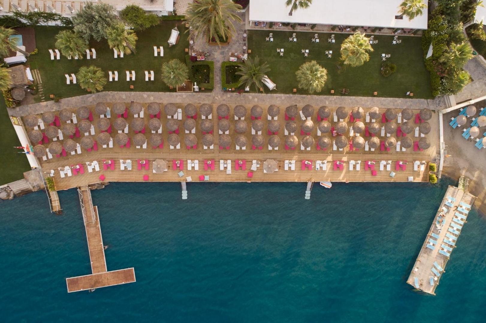 Prive Hotel Bodrum aerofoto uz koka sauļošanās platformu pie jūras ar rozā akcentiem, saulessargiem un zālienu ar palmām.