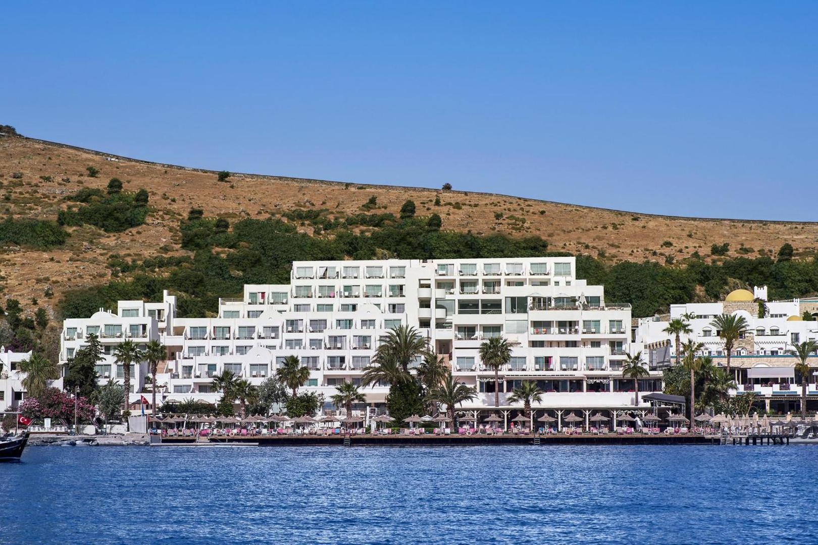 Prive Hotel Bodrum baltā kaskādes tipa ēka skatā no jūras puses ar palmām un kalnu fonā saulainā dienā.