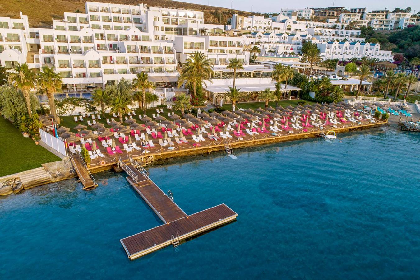 Prive Hotel Bodrum skats no putna lidojuma uz viesnīcas balto ēku, koka piestātni un sauļošanās zonu pie dzidri zilā jūras ūdens Bodrumā.