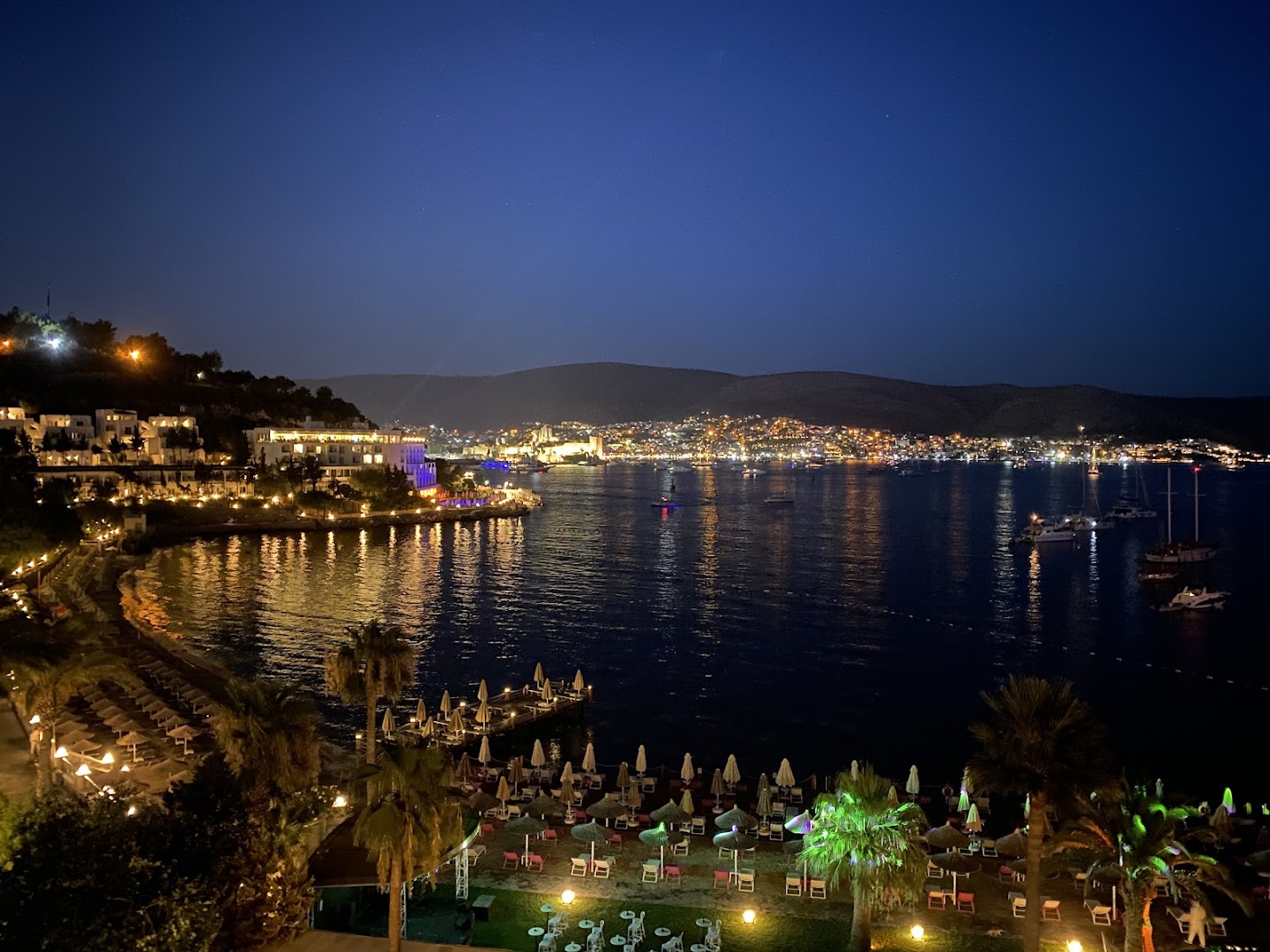 Prive Hotel Bodrum skats naktī uz izgaismoto Bodrumas pilsētu un līci no viesnīcas piekrastes zonas ar palmām un saulessargiem.