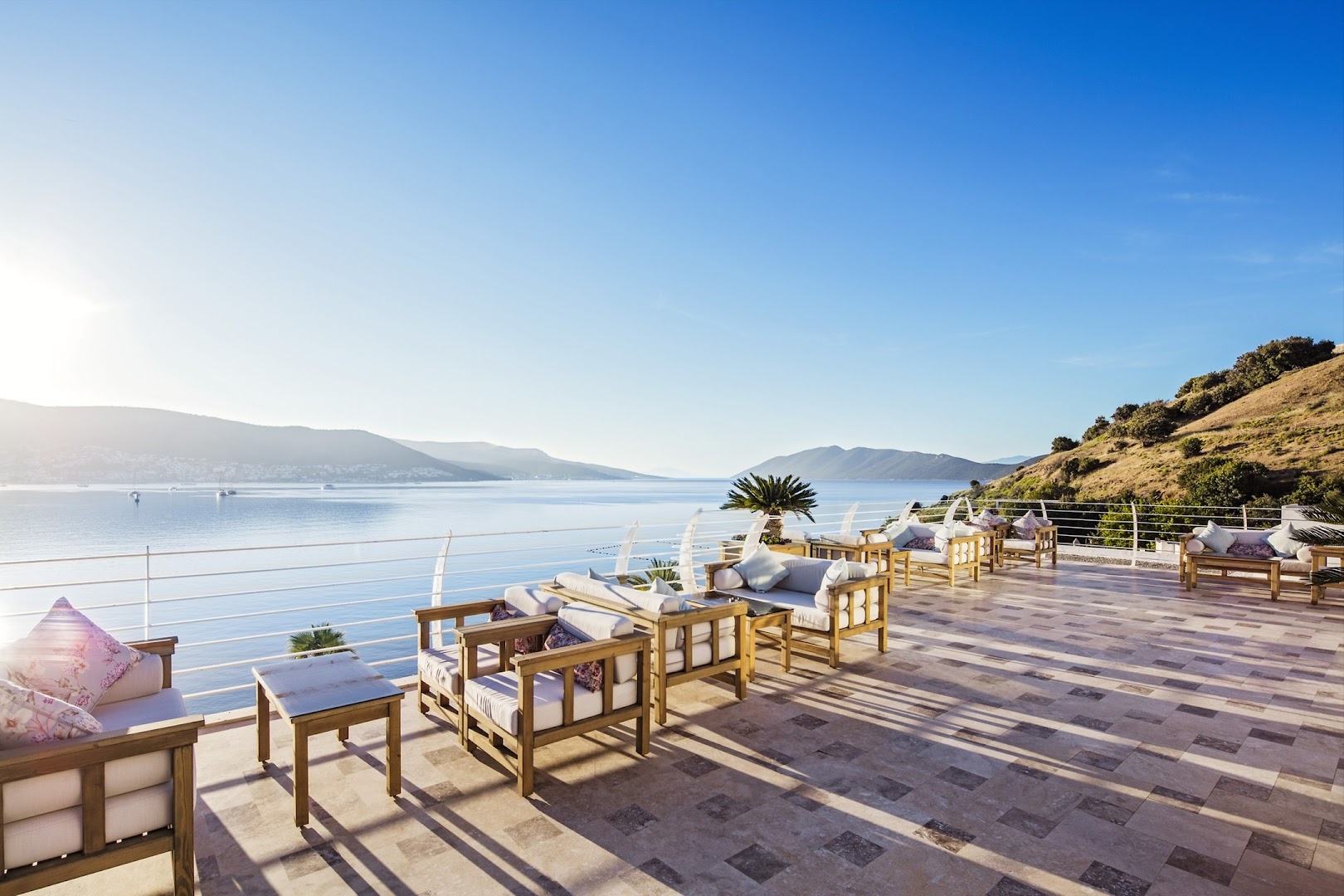 Prive Hotel Bodrum saulaina āra terase ar koka mēbelēm, mīkstiem dīvāniem un panorāmas skatu uz jūru un kalniem saulrietā.