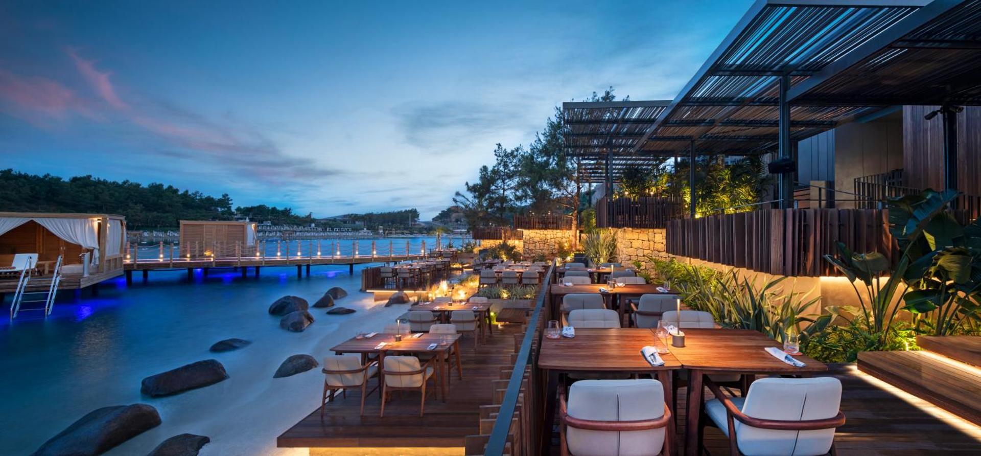 Lujo Hotel Bodrum restorāna terase vakarā pie jūras ar koka klāju, elegantiem galdiem un skatu uz izgaismotu piestātni