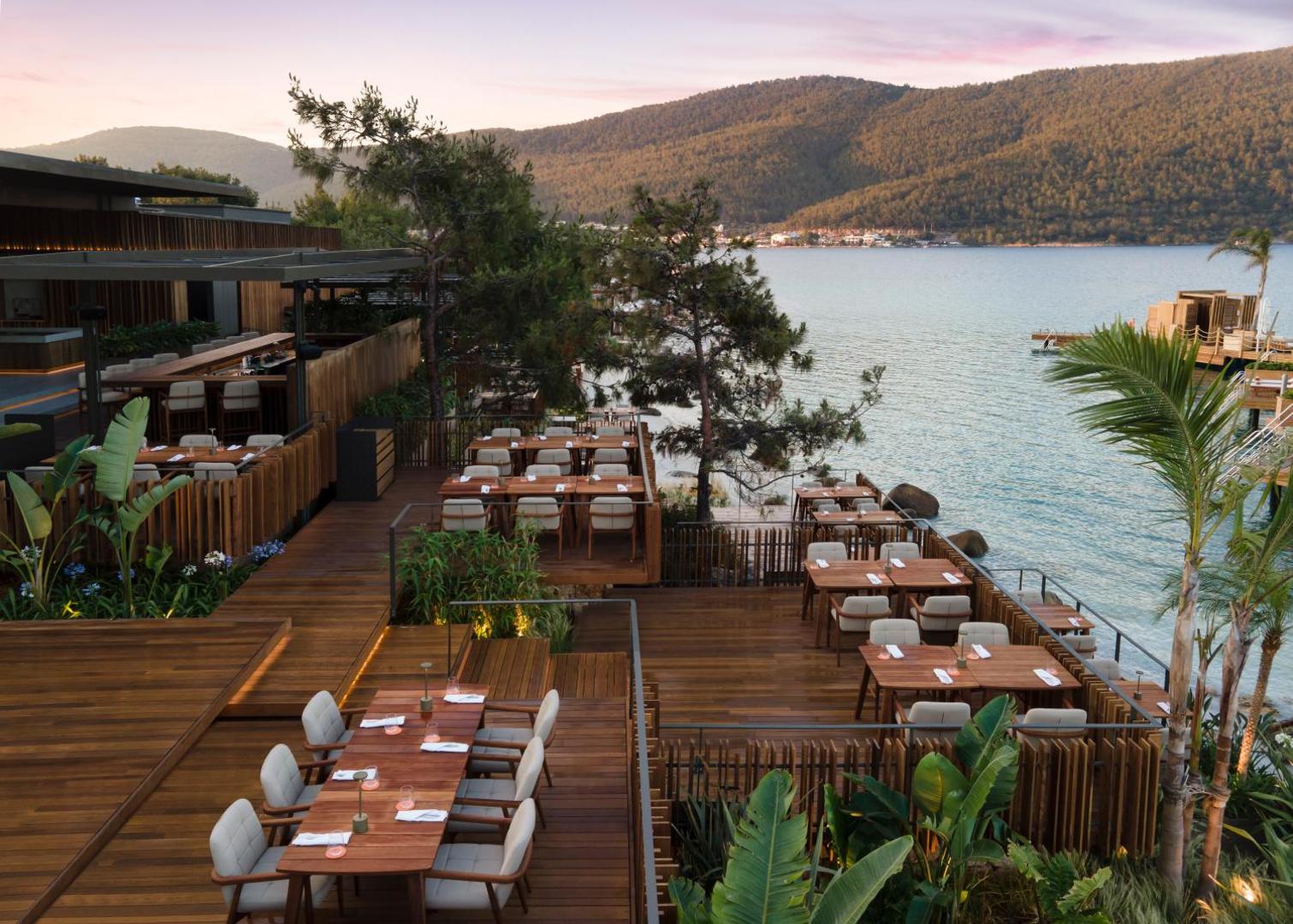 Lujo Hotel Bodrum restorāna terase dienas laikā ar skatu uz Egejas jūras līci un kalniem, koka mēbelēm un palmām