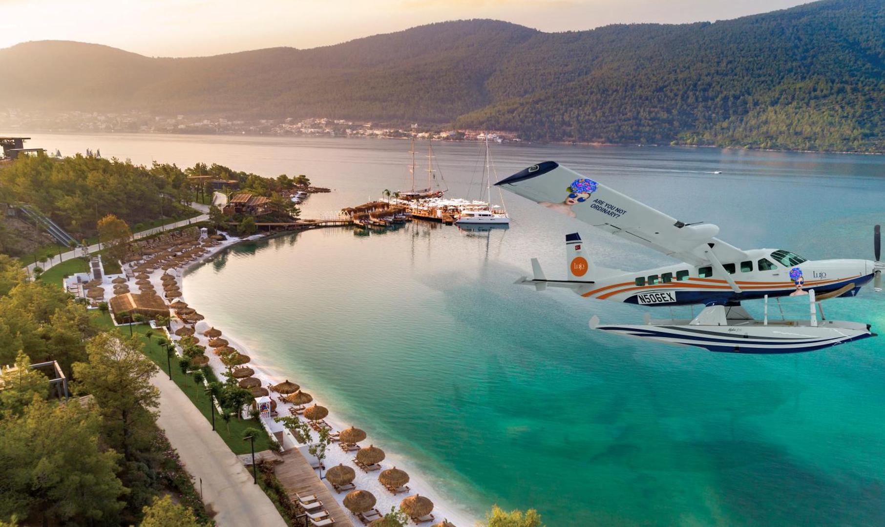 Lujo Hotel Bodrum privātā pludmale ar baltām smiltīm, saulessargiem un viesnīcas hidroplānu virs tirkīzzila Egejas jūras līča