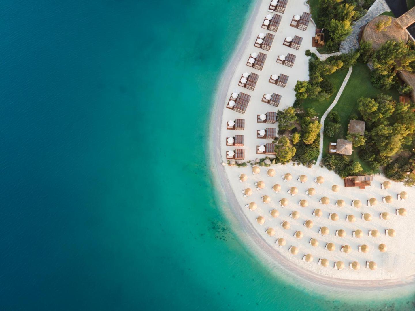 Lujo Hotel Bodrum privātā pludmale no putna lidojuma ar saulessargiem, sauļošanās krēsliem un dzidru, tirkīzzilu ūdeni Bodrumas piekrastē.