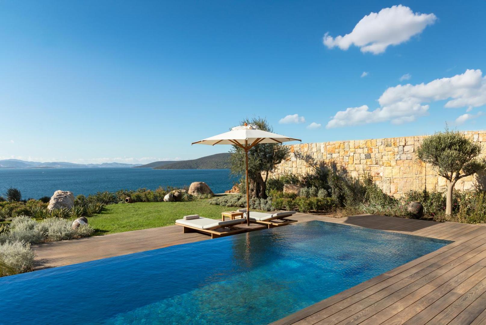 Lujo Hotel Bodrum privātais bezmalas baseins ar skatu uz Egejas jūru, koka terasi, sauļošanās krēsliem un saulessargu piekrastē.