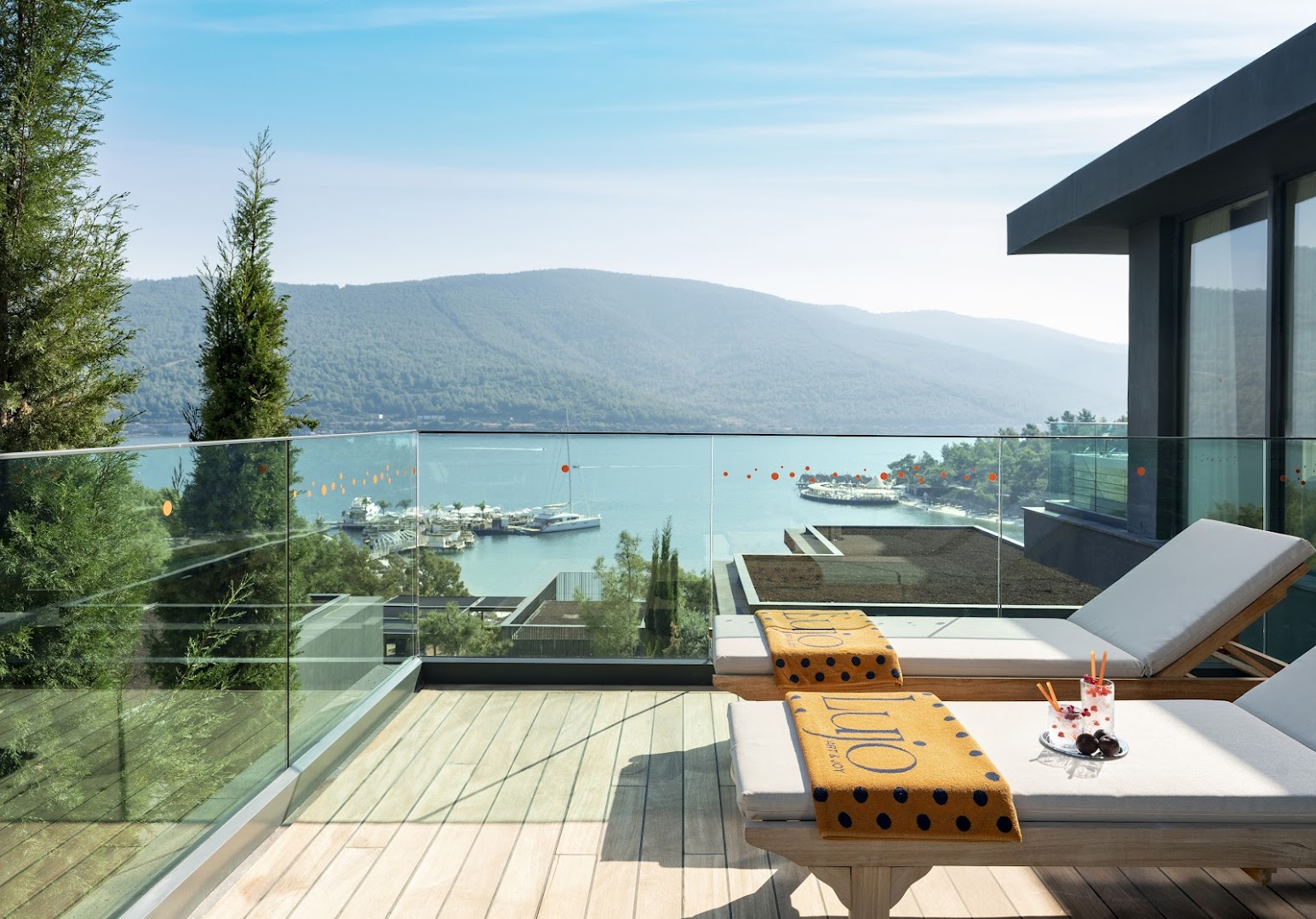 Lujo Hotel Bodrum terase ar sauļošanās krēsliem, kokteiļiem un skatu uz līci, jahtām un kalniem tālumā