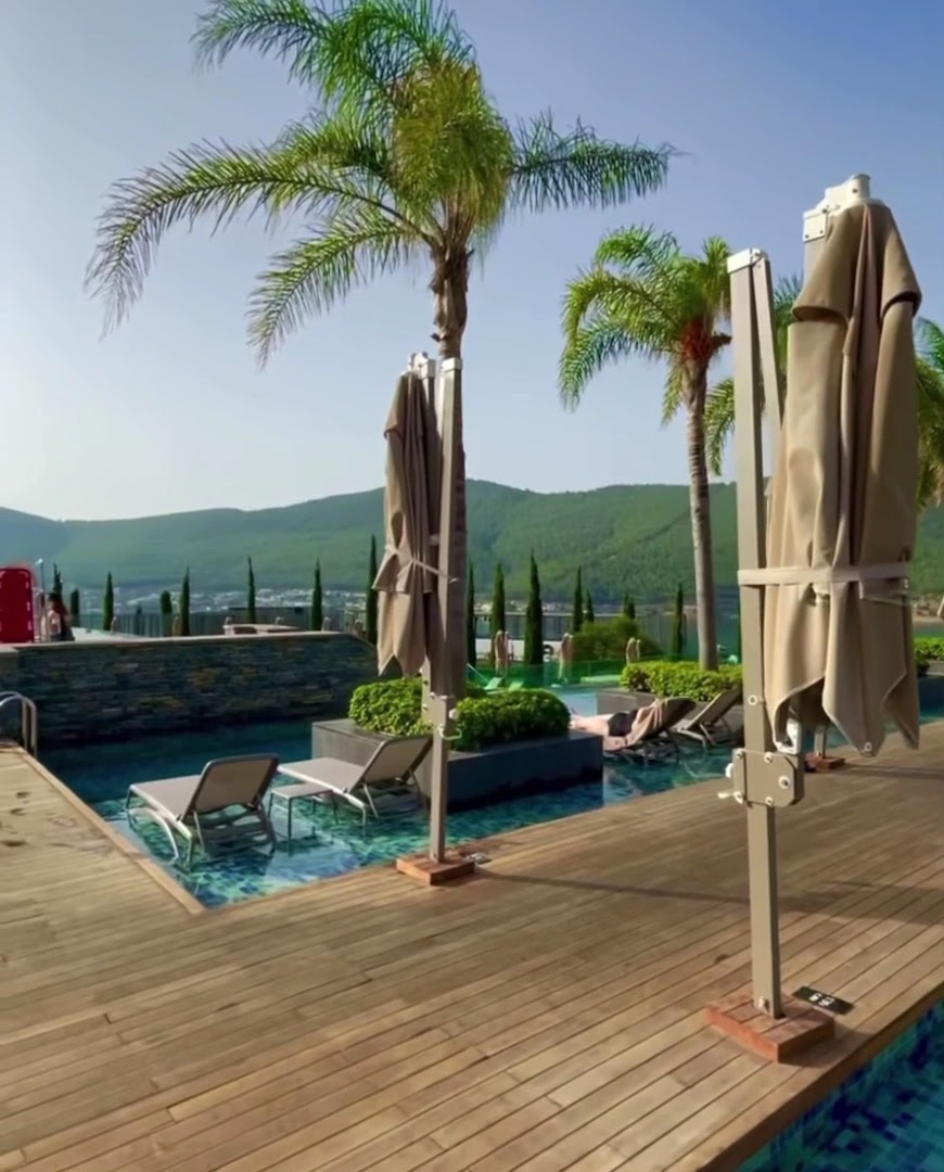 Lujo Hotel Bodrum āra baseina zona ar palmām, sauļošanās krēsliem ūdenī un skatu uz kalniem Bodrumas piekrastē.