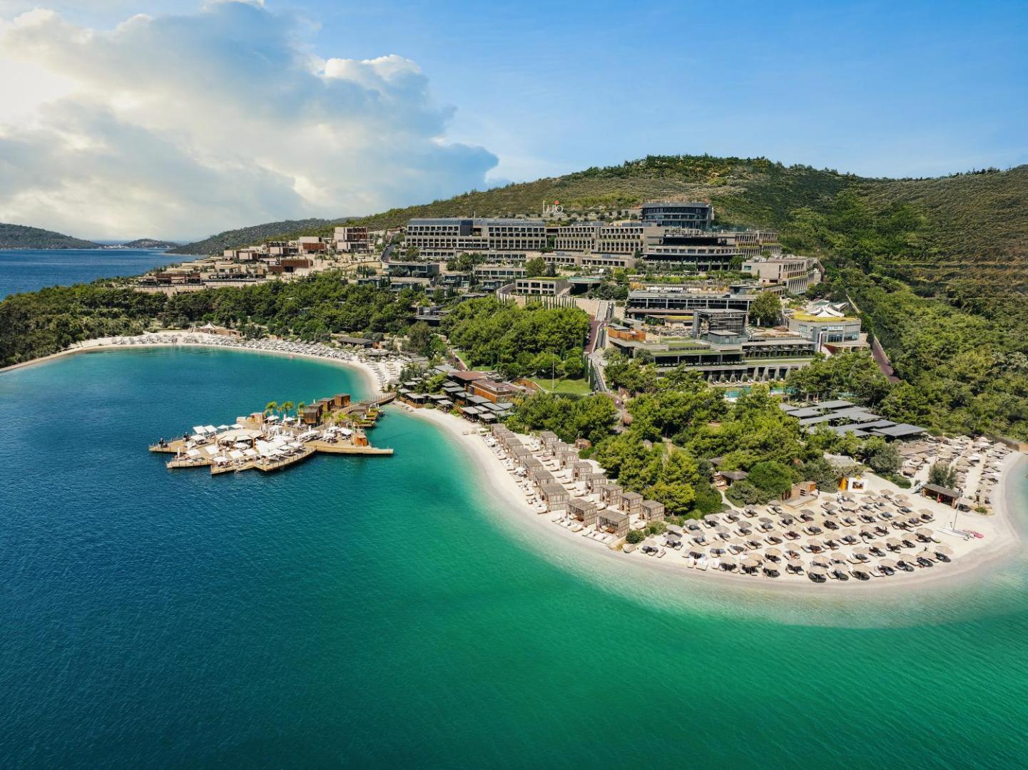 Lujo Hotel Bodrum panorāmas skats uz viesnīcas kompleksu kalna nogāzē pie tirkīzzila līča ar privātām pludmalēm un zaļojošu apkārtni
