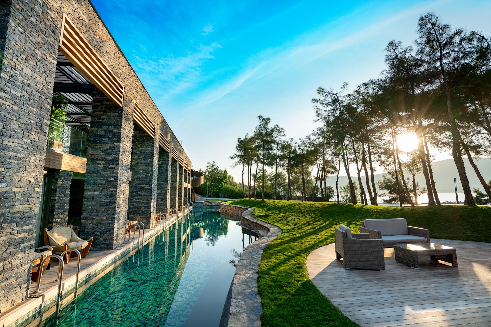 Lujo Hotel Bodrum modernā arhitektūra ar akmens fasādi, garu peldbaseinu gar ēku un atpūtas zonu priežu ielokā saulrietā.