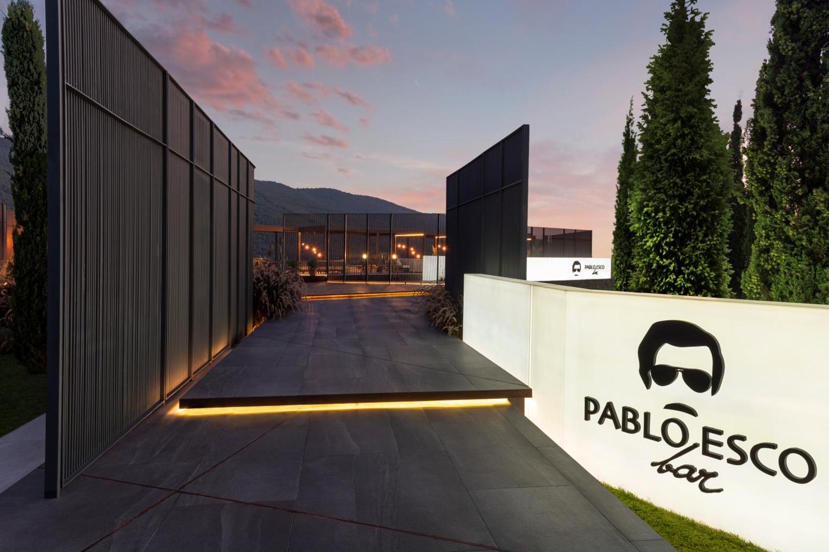 Lujo Hotel Bodrum Pablo Esco bāra ieeja saulrieta laikā ar modernu apgaismojumu, tumšām dizaina sienām un ciprešu kokiem