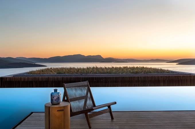 Lujo Hotel Bodrum terase ar koka krēslu pie bezmalas baseina saulrieta laikā ar skatu uz jūras līci un salām.