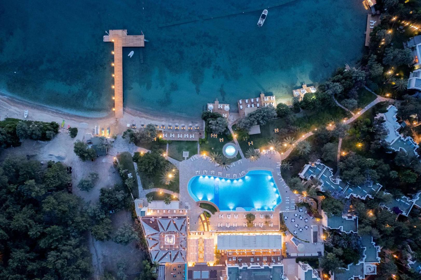 DoubleTree by Hilton Bodrum Isil Club Resort vakara skats no augšas uz izgaismotu baseinu, terasēm un koka piestātni pie tumšajiem jūras ūdeņiem.