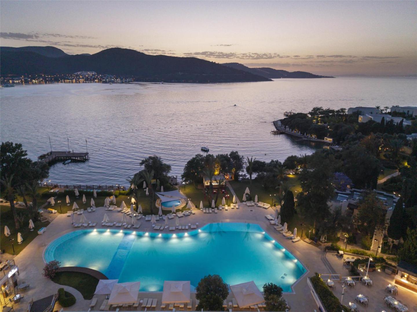 DoubleTree by Hilton Bodrum Isil Club Resort skats no augšas vakarā — izgaismots baseins, piekrastes līnija un kalni saulrietā