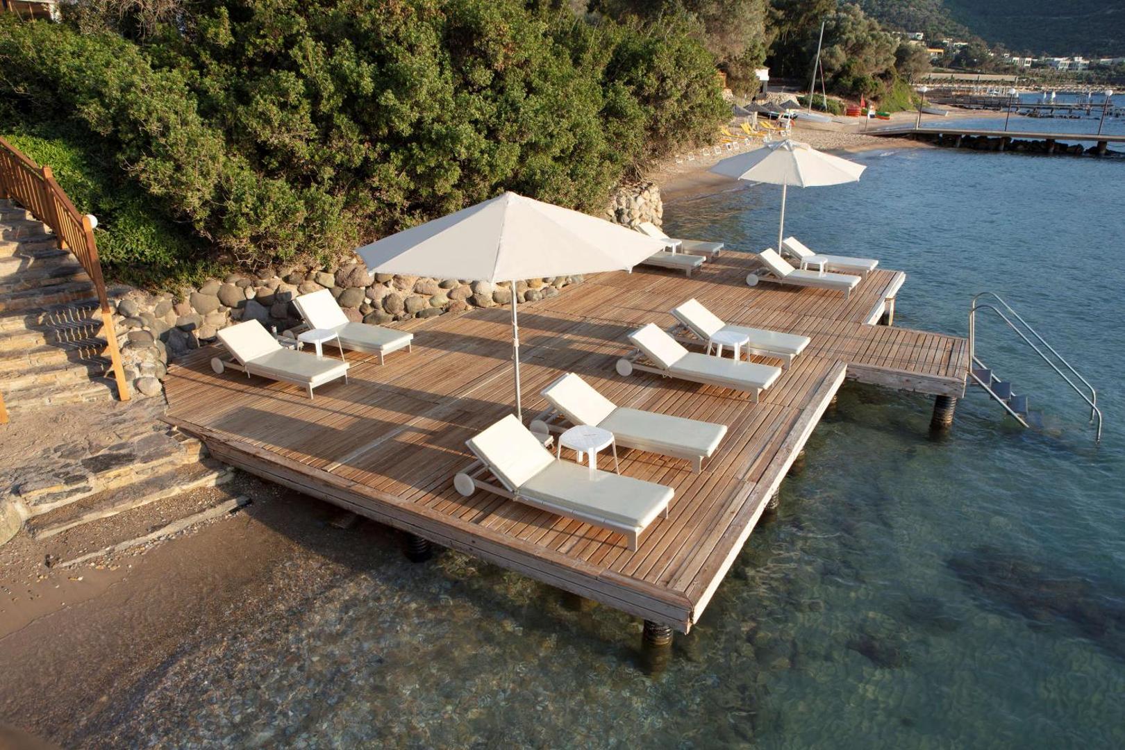 DoubleTree by Hilton Bodrum Isil Club Resort koka sauļošanās platforma virs ūdens — balti zviļņi un saulessargi pie dzidras jūras