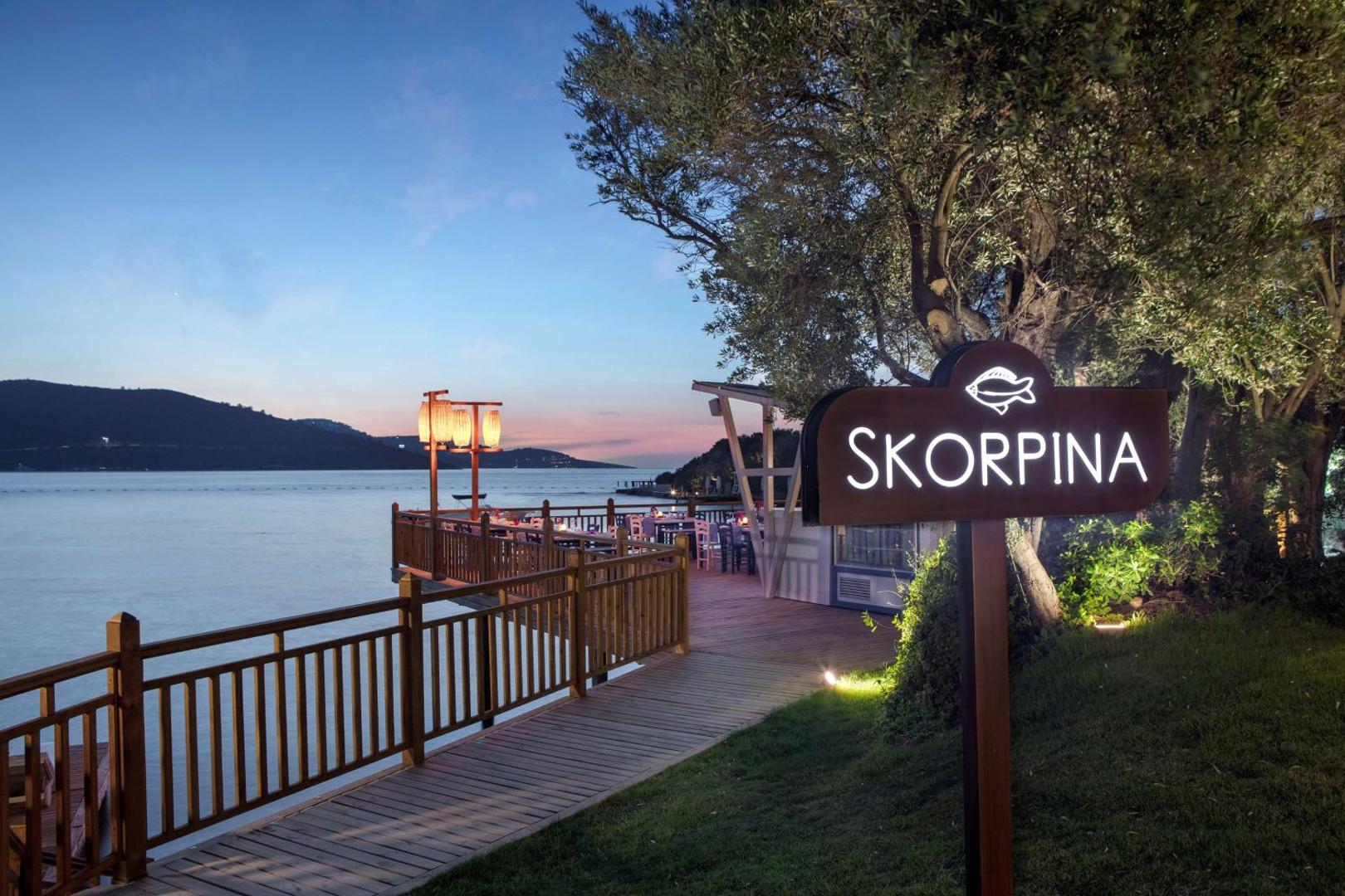 DoubleTree by Hilton Bodrum Isil Club Resort restorāna Skorpina ieeja un koka laipa saulrietā — skats uz līci un kalniem tālumā