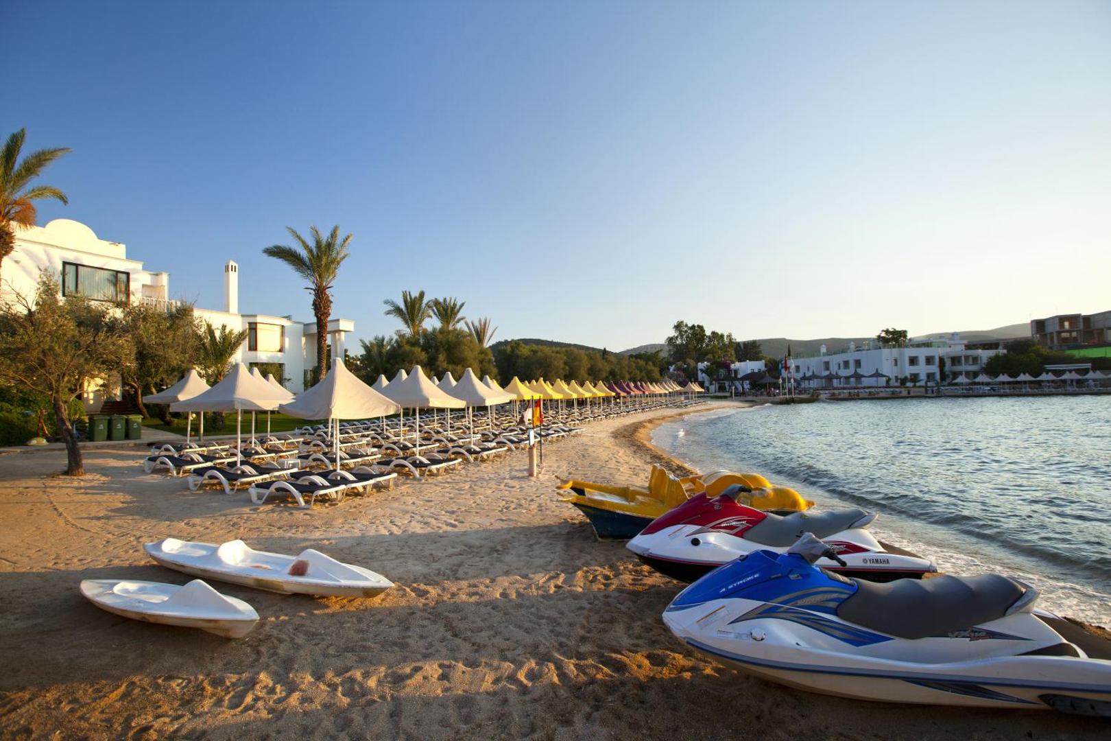 Samara Hotel Bodrum privātā smilšu pludmale ar saulessargiem, sauļošanās krēsliem un ūdens motocikliem pie dzidrā Egejas jūras krasta